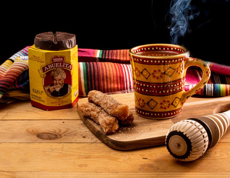 Mexican hot chocolate with churros on a wooden board, served with a molinillo and colorful blanket.